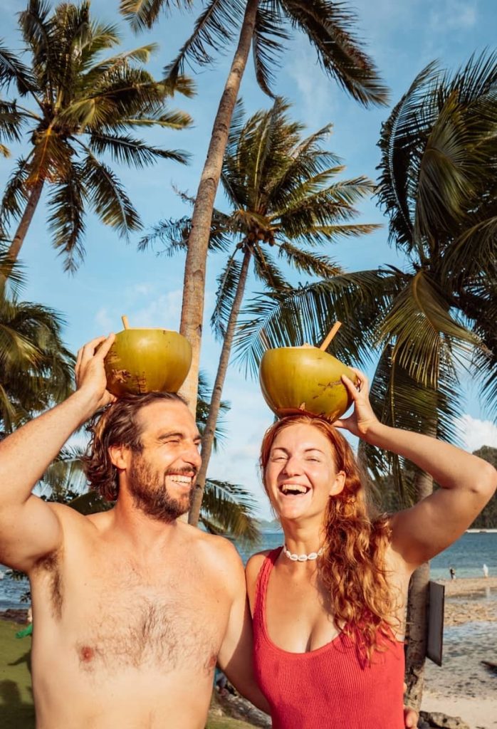 A couple smiles and balances coconuts on their heads under palm trees at the beach in Siargao, Philippines.