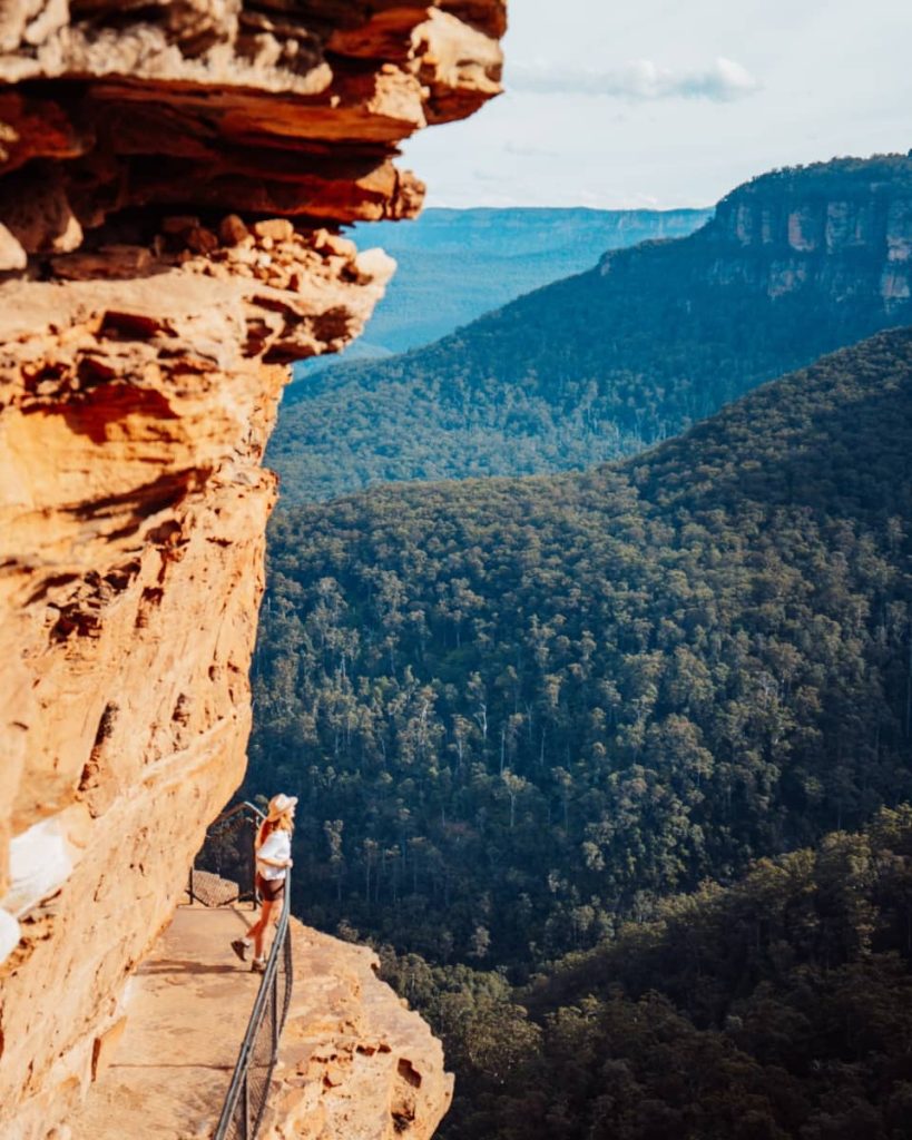 Woman standing on a narrow cliffside path with a railing, overlooking the forested valley near Wentworth Falls in the Blue Mountains, NSW.