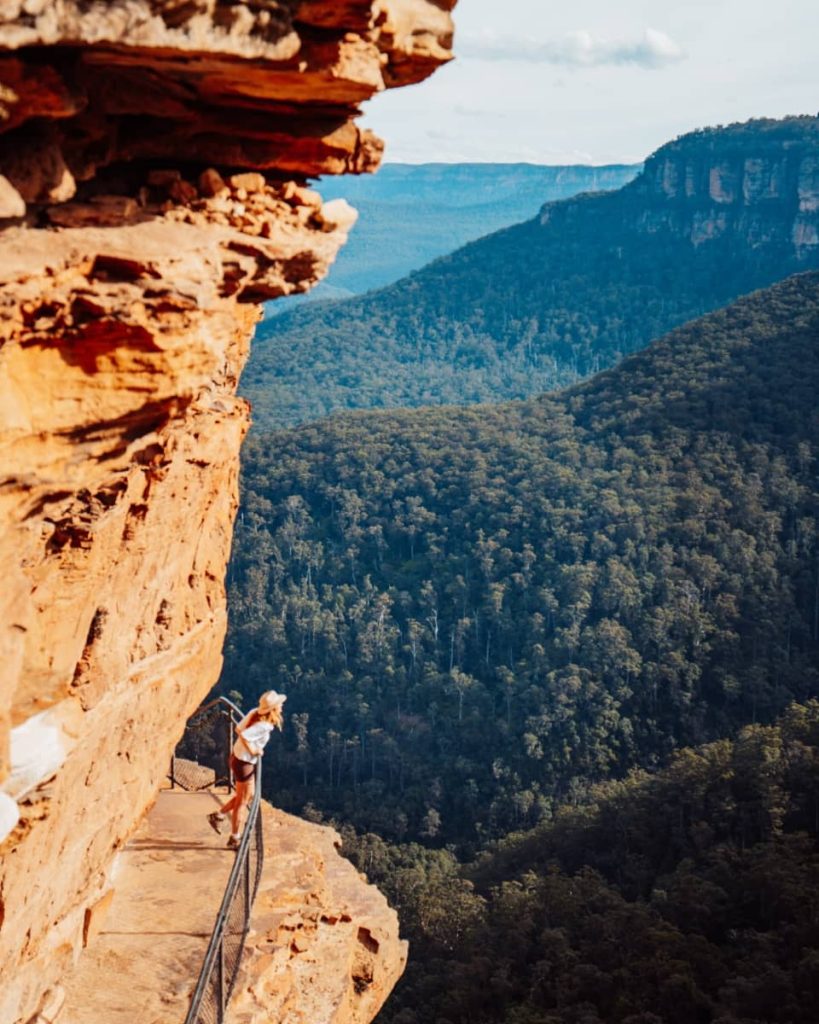 A hiker standing on a narrow cliffside path overlooking dense forest and mountain ridges in the Blue Mountains near Wentworth Falls, NSW.