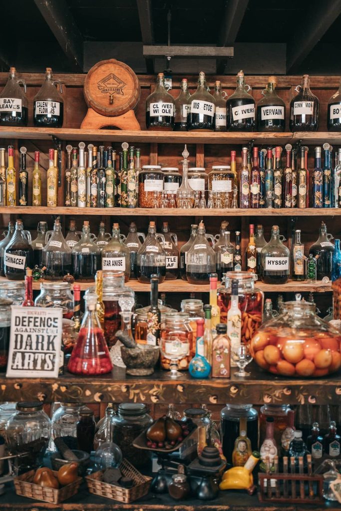 A wooden apothecary-style display filled with labeled glass jars, vintage bottles, and potion-like concoctions. Shelves are stocked with herbs, spices, and tinctures such as "star anise," "mistletoe," and "cloves." A sign reading "Defence Against the Dark Arts" adds a magical touch to the rustic, mystical atmosphere.