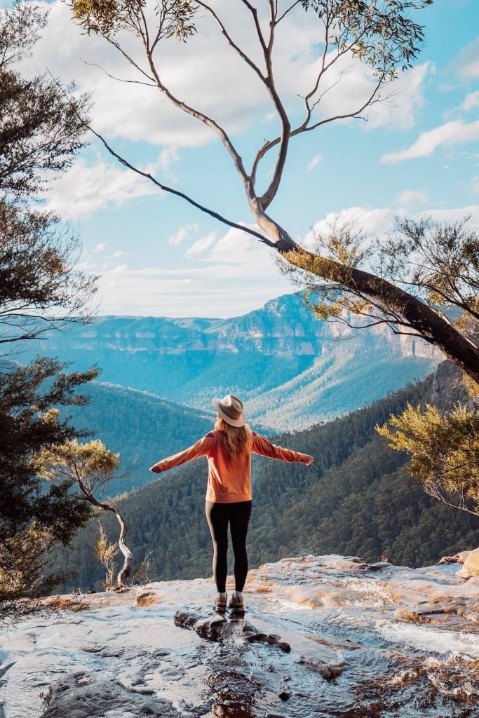 Woman standing on wet rock near the edge of Fortress Falls, arms outstretched, overlooking forested valleys and cliffs in the Blue Mountains, NSW.