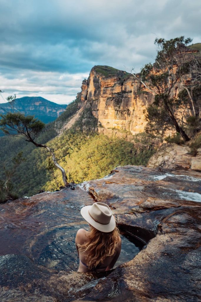 Woman in a hat sitting in a natural rock infinity pool at Fortress Falls, gazing over the escarpment and forested valleys of the Blue Mountains.