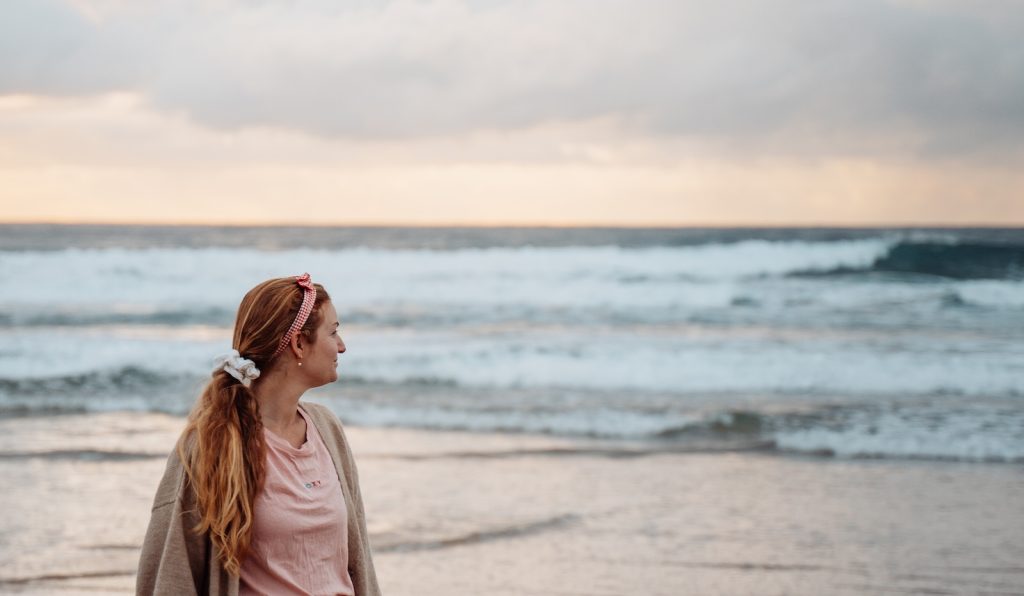 A woman stands on the shoreline of Gold Coast Broadwater, gazing out at the rolling waves under a cloudy sky. She wears a pink shirt, a light cardigan, and a headband, her hair gently tousled by the ocean breeze. The horizon glows faintly with the last light of the day, reflecting on the water.