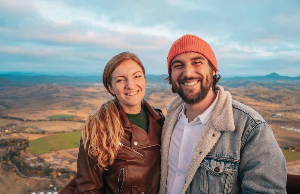 A smiling couple poses for a photo high above the landscape during a hot air balloon ride over the Gold Coast hinterland. The woman wears a brown leather jacket over a green sweater, while the man wears a denim jacket with a sherpa lining and a red beanie. Rolling hills, farmland, and a distant lake stretch behind them under a soft morning sky.