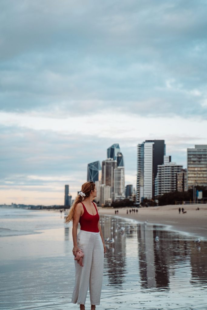 A woman in a red swimsuit and white pants stands on a wet sandy beach, looking towards the Gold Coast skyline. The reflective shoreline mirrors the tall modern buildings, while seagulls scatter along the water’s edge. The sky is overcast, creating a soft, moody atmosphere.