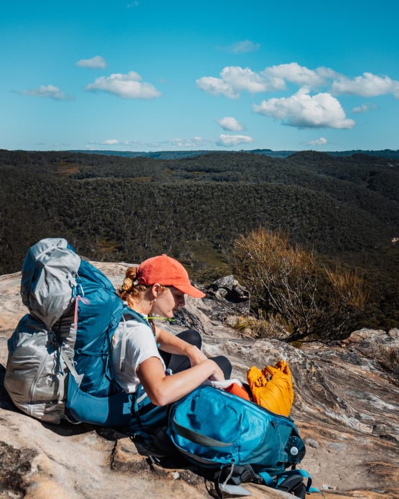 Woman sitting on a rocky outcrop in the Blue Mountains with a large hiking pack, surrounded by bushland under a blue sky.