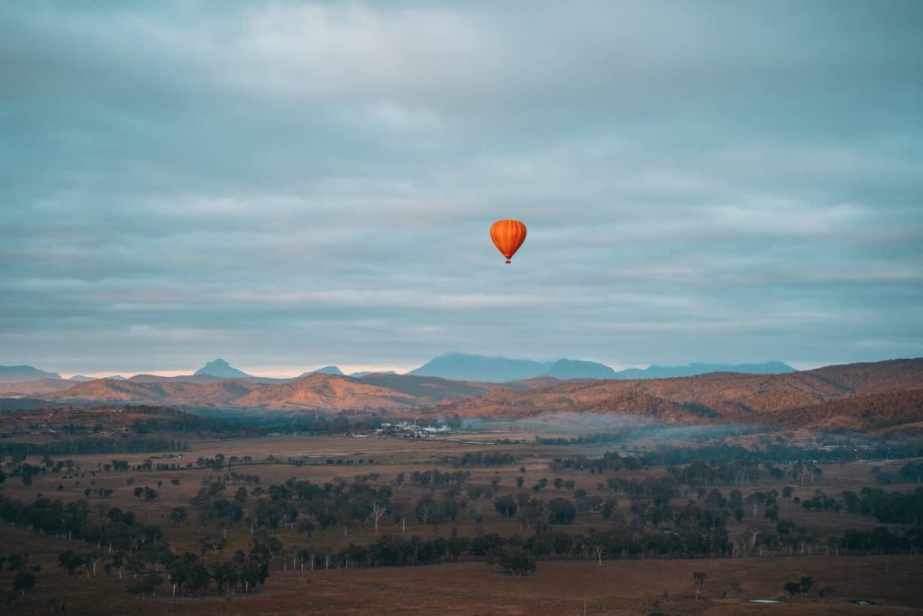 A single vibrant orange hot air balloon floats above the rolling countryside of the Gold Coast hinterland. The vast landscape stretches towards distant mountains, with golden morning light casting soft shadows on the terrain. A thin layer of mist lingers in the valley, adding to the peaceful atmosphere.