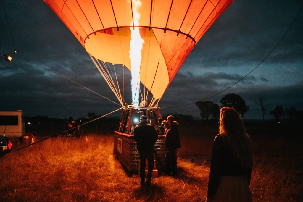 A hot air balloon is being inflated with a bright flame before sunrise in an open field near the Gold Coast, Australia. A group of people stands around the basket, preparing for takeoff, while a woman in the foreground watches the process. The dark sky and glowing balloon contrast, creating a dramatic early morning scene.