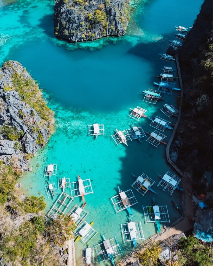 Aerial view of traditional Filipino outrigger boats (bangkas) anchored in the crystal-clear turquoise waters near the entrance to Kayangan Lake, surrounded by towering limestone cliffs in Coron, Palawan.