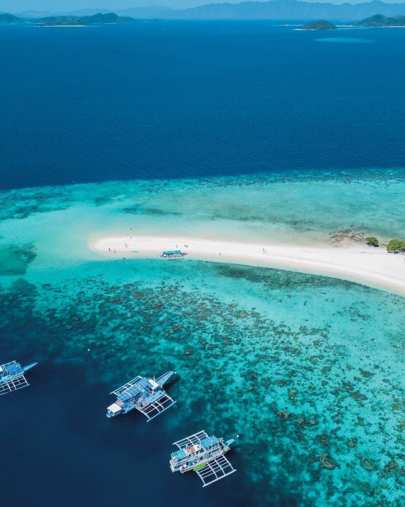 Aerial view of traditional Filipino outrigger boats anchored in turquoise water near a narrow white sandbar surrounded by coral reef, with deep blue ocean and distant islands in the background.