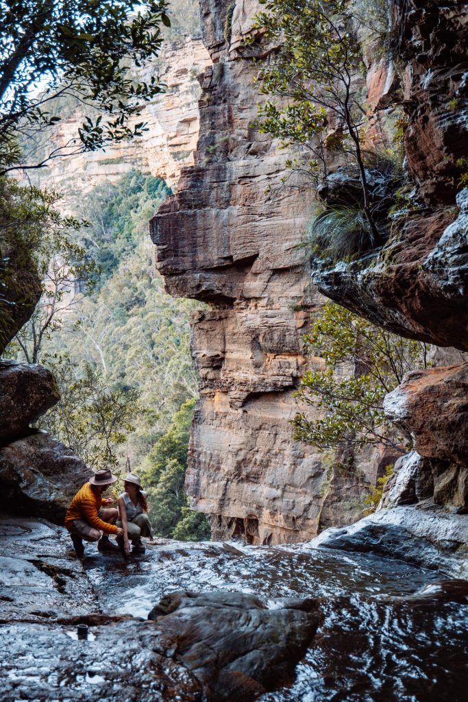 Two people crouched beside the edge of a rock pool at Leura Cascades, surrounded by towering cliffs and forest in the Blue Mountains, NSW.