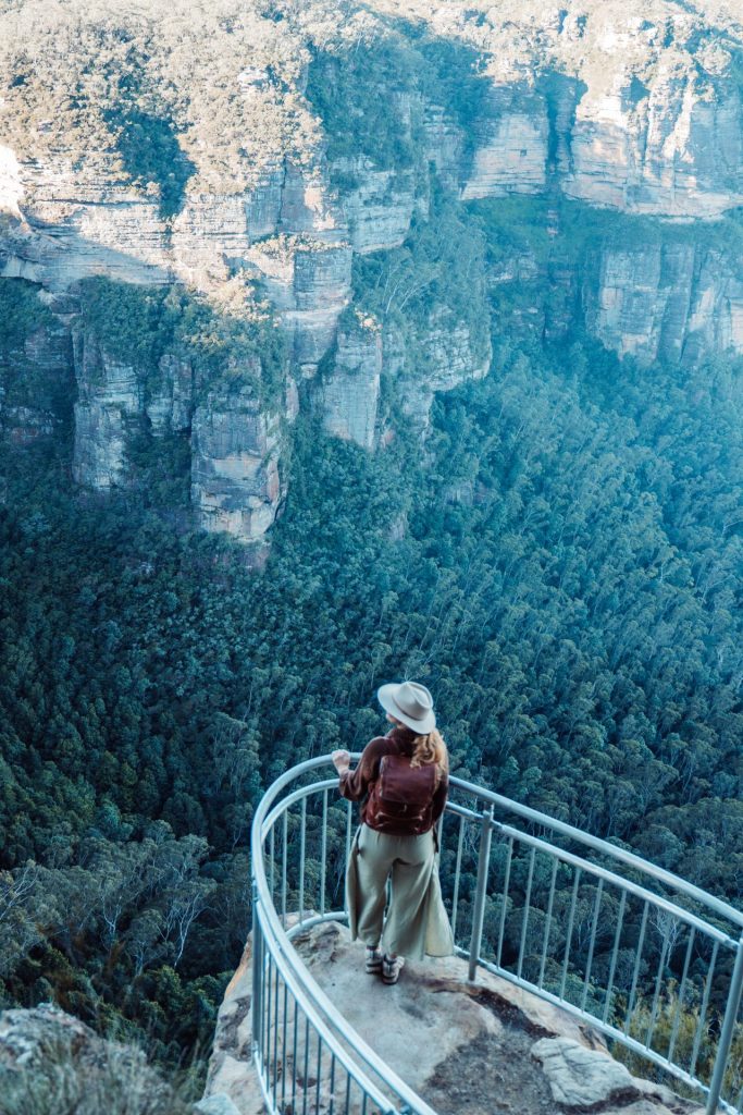 Woman standing at a cliffside lookout with guardrails, overlooking a forested valley and dramatic cliffs near Leura Cascades in the Blue Mountains, NSW.