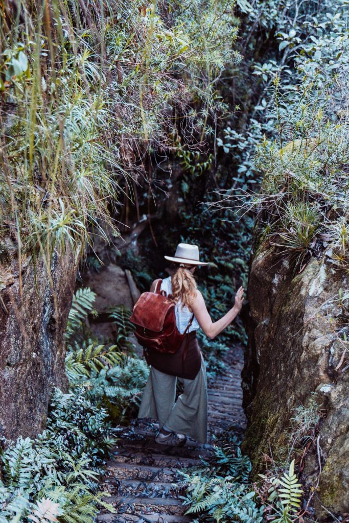 Woman with a backpack walking down a narrow fern-lined trail between rocky cliffs near Leura Cascades in the Blue Mountains, NSW.