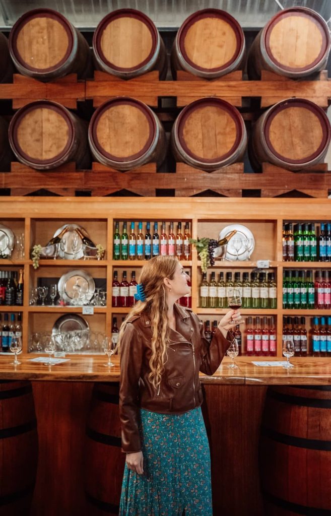 A woman in a brown leather jacket holds a glass of red wine while admiring the wooden wine barrels stacked above the bar at a Mount Tamborine winery. Behind her, shelves are lined with colorful bottles of wine, neatly arranged, alongside polished glassware. The cozy setting highlights the charm of a boutique wine-tasting experience in the Gold Coast hinterland.
