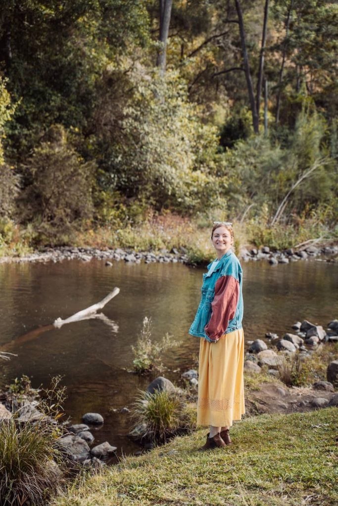 A woman stands by the edge of a calm river, smiling at the camera, surrounded by lush greenery. She wears a colorful patchwork denim jacket over a flowing yellow skirt, blending into the natural landscape. The riverbank is lined with rocks and grass, with a fallen tree branch partially submerged in the water.