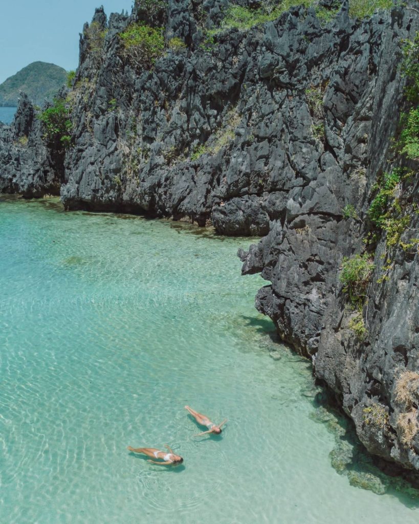 Two women floating in crystal-clear turquoise water next to jagged limestone cliffs in a secluded lagoon in El Nido, Philippines.