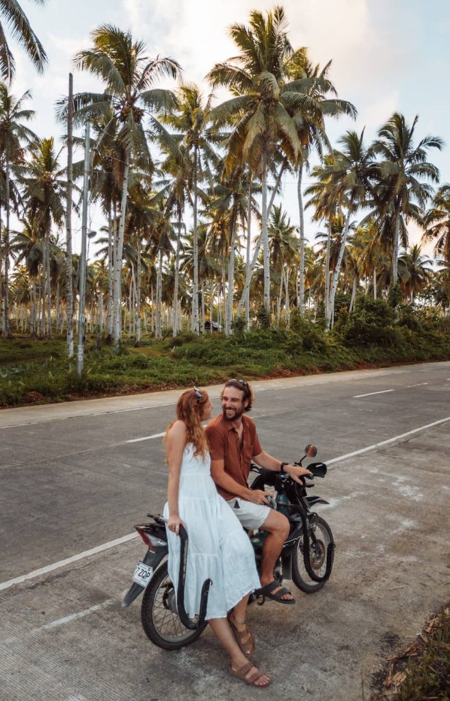 A couple sits on a motorbike beside a palm-lined road in Siargao, Philippines, smiling at each other at golden hour.
