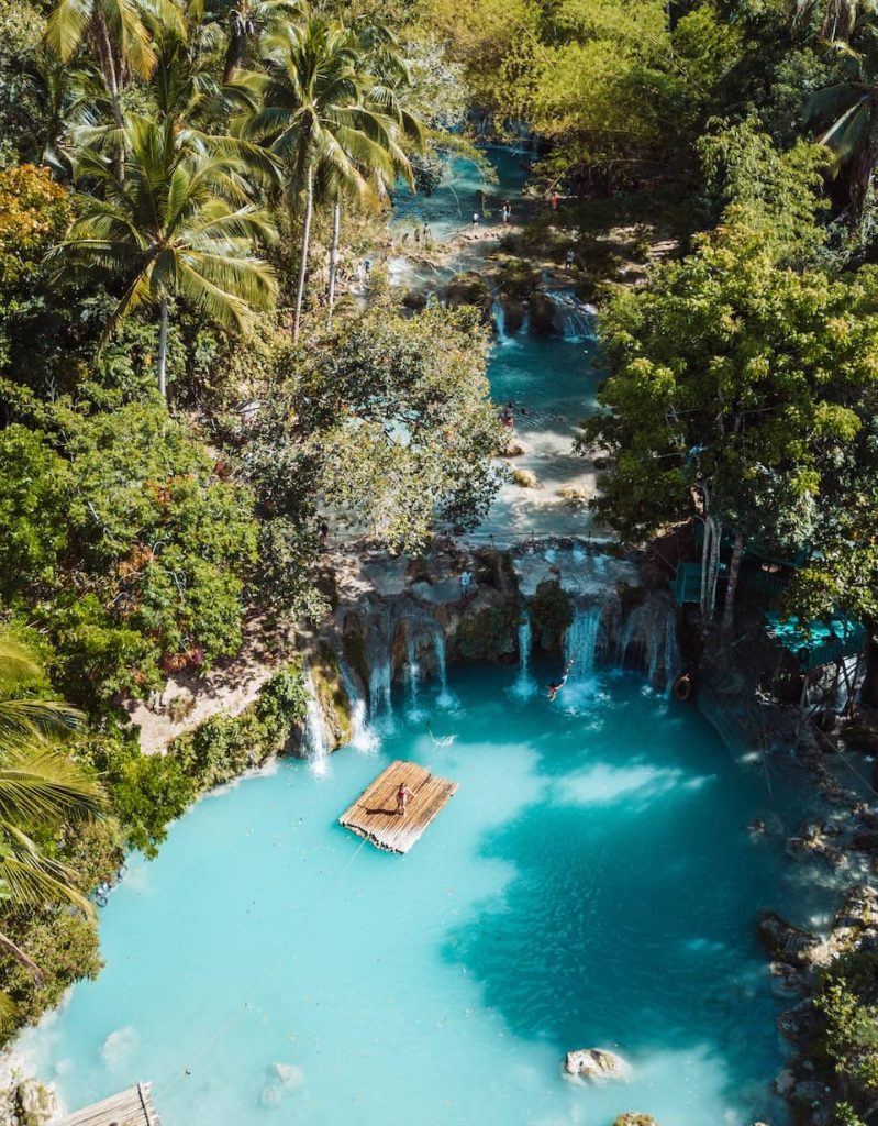 Aerial view of Cambugahay Falls in Siquijor, Philippines, with turquoise blue water, lush jungle, and a bamboo raft floating below the waterfalls.