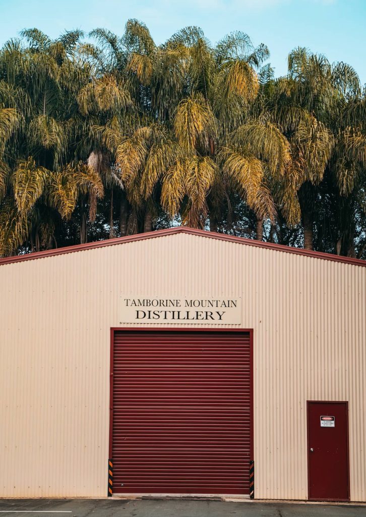 A beige metal building with a red roller door and a sign reading "Tamborine Mountain Distillery" stands against a backdrop of lush green palm trees. The simple structure contrasts with the dense foliage, giving a rustic yet tropical feel.