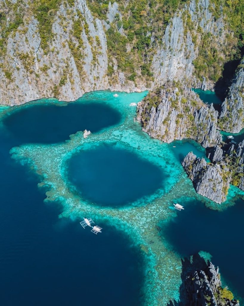 Aerial view of the Twin Lagoon in Coron, Palawan, with turquoise waters, dark blue pools, and dramatic limestone cliffs rising from the sea. Traditional Filipino boats float nearby.