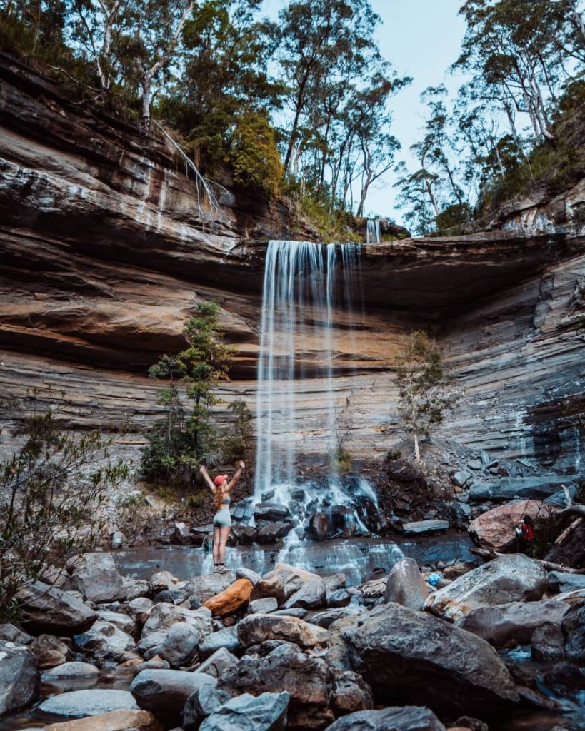 Woman standing on rocks with arms raised beneath the flow of Victoria Falls, surrounded by sandstone cliffs and bush in the Blue Mountains.