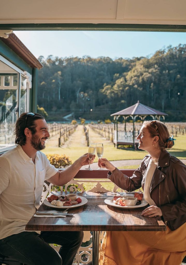 A couple toasts with glasses of sparkling wine while enjoying a breakfast spread at an outdoor table overlooking a vineyard. The sun casts a golden glow over the scene, highlighting the lush greenery and a charming gazebo in the background.