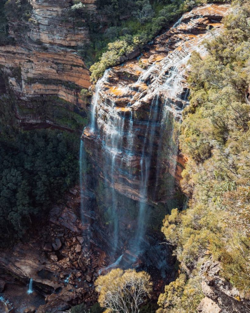 Aerial view of Wentworth Falls pouring down a tall sandstone cliff in the Blue Mountains, surrounded by rugged bushland and forest.