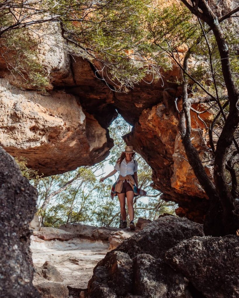 Woman walking through a natural rock arch surrounded by bushland, along a trail near Wentworth Falls in the Blue Mountains, NSW.