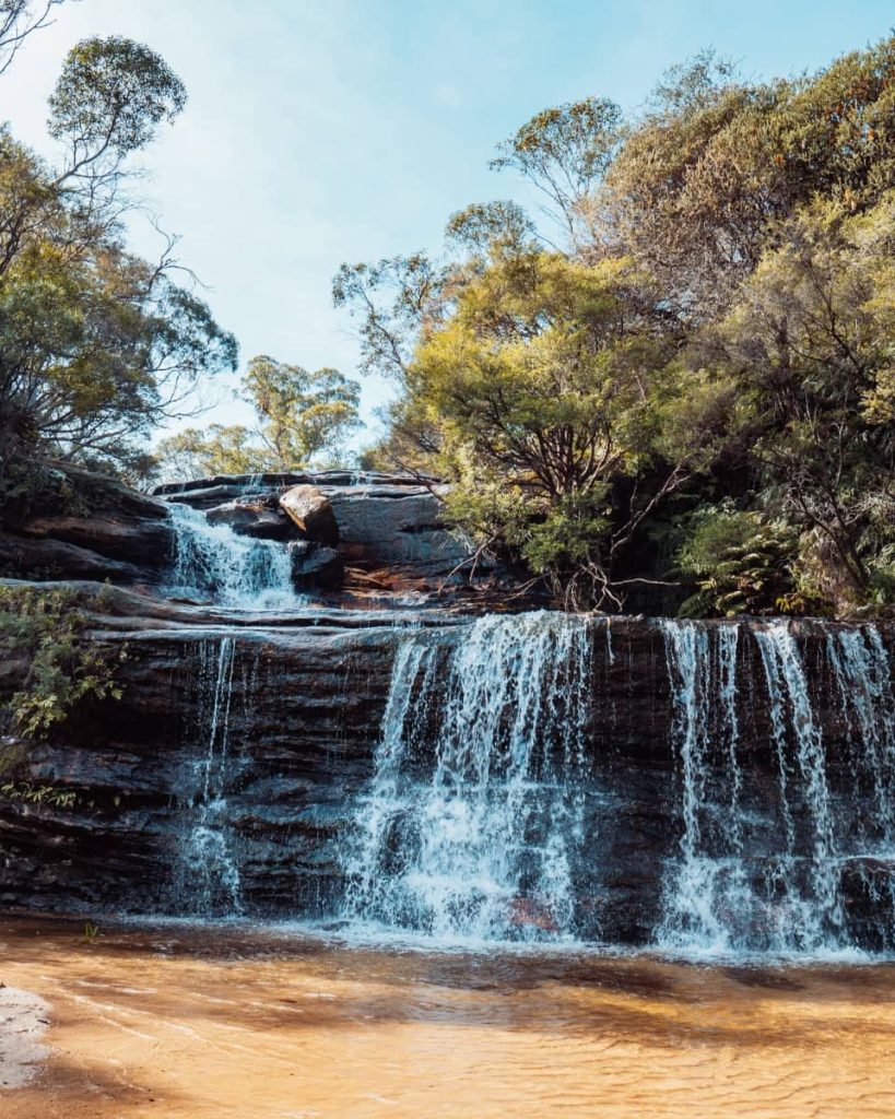 A wide-angle view of a tiered waterfall cascading over dark rock ledges, surrounded by bushland in the Blue Mountains region of NSW.