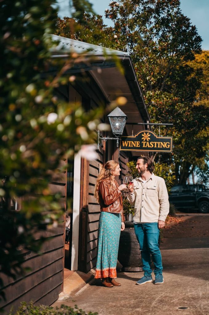 A couple stands outside a rustic wooden winery on Mount Tamborine, Gold Coast, Australia, clinking glasses of red wine. The woman wears a brown leather jacket over a flowing blue dress, while the man is dressed casually in jeans and a light-colored shirt. A "Wine Tasting" sign hangs above them, and the sun casts a warm glow on the scene.