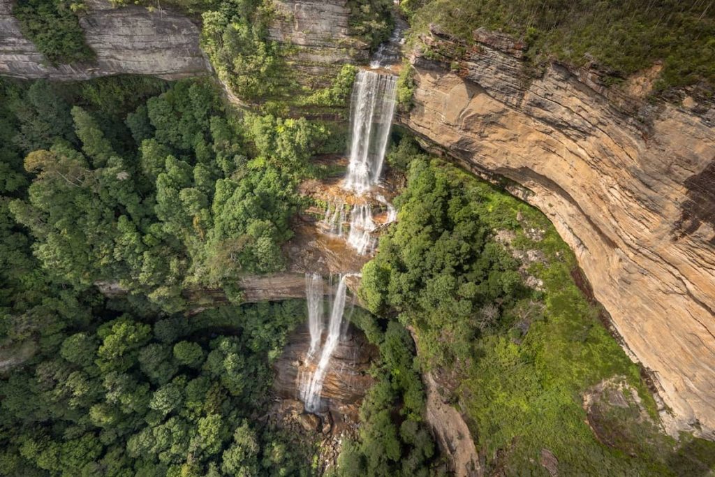 Aerial view of Katoomba Falls cascading in multiple stages down a cliffside surrounded by lush forest in the Blue Mountains, Australia.