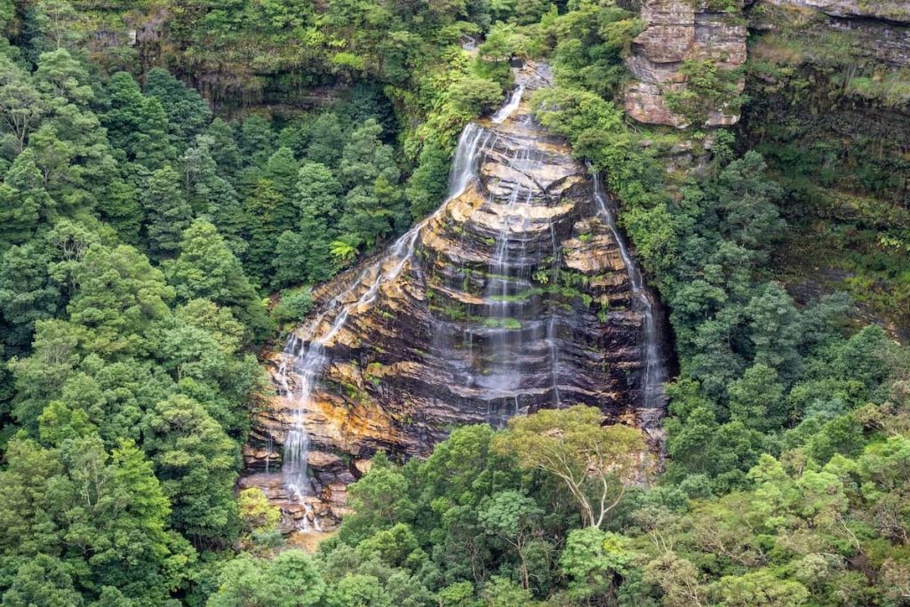 Aerial view of Bridal Veil Falls cascading down terraced rock layers and surrounded by dense green forest in the Blue Mountains, Australia.