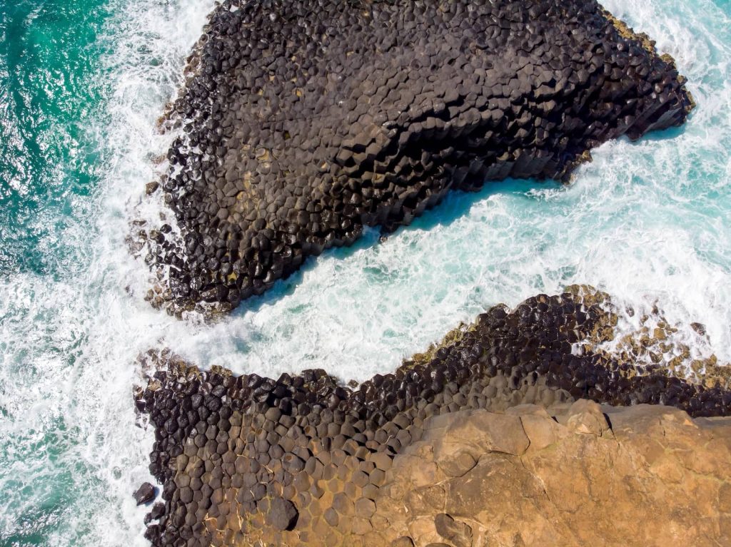 Aerial view of the unique hexagonal basalt columns at Fingal Head in the Gold Coast, Australia. The dark volcanic rock formations extend into the turquoise ocean, with white waves crashing between the separated sections of rock. The contrast between the structured columns and the swirling sea highlights the rugged beauty of this coastal landmark.