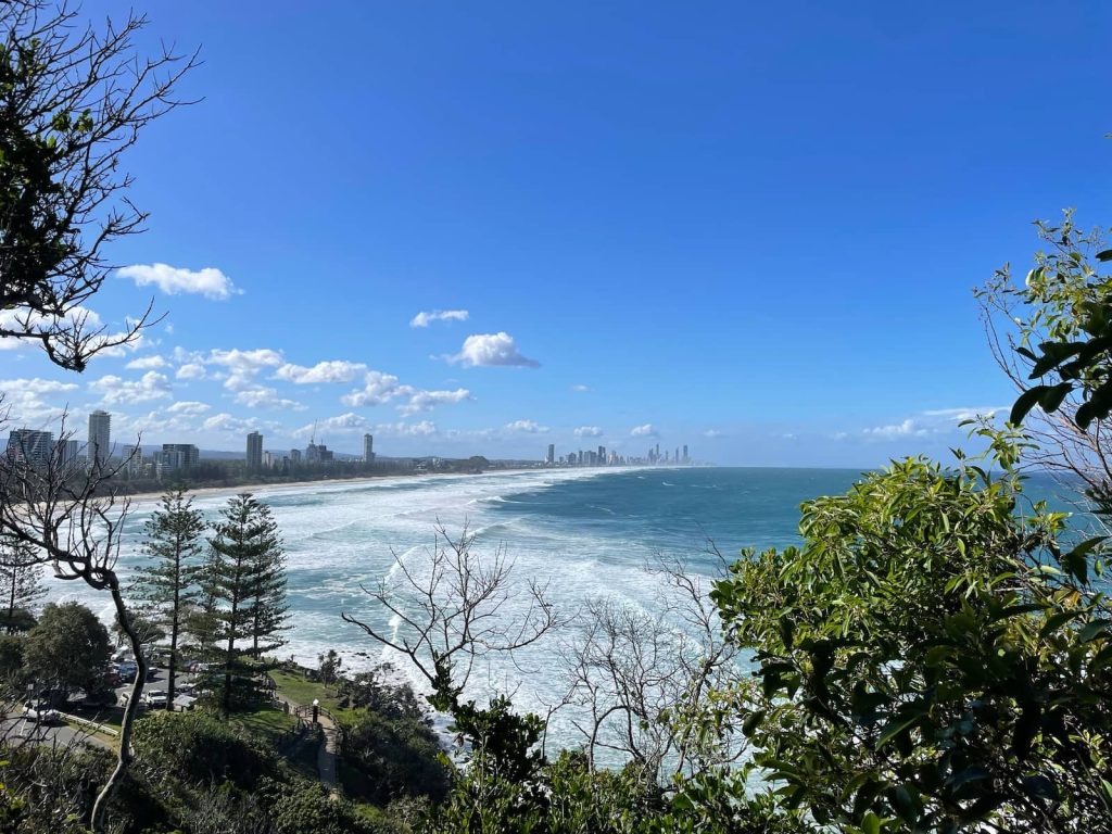 A panoramic view of the Gold Coast, Queensland, featuring a blue ocean with rolling waves, a sandy shoreline lined with high-rise buildings, and green coastal vegetation framing the scene. The skyline of Surfers Paradise rises in the distance under a bright blue sky with scattered clouds.