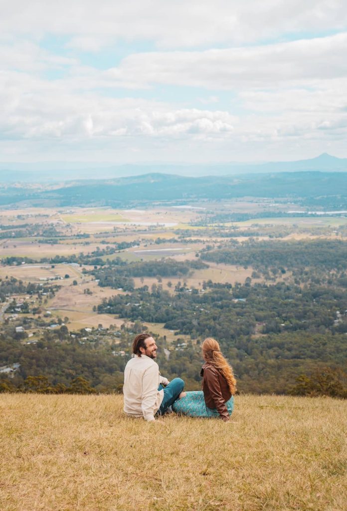 A couple sits on a grassy hilltop, overlooking a vast valley with scattered houses, trees, and distant mountain ranges. The man, wearing a light-colored shirt, leans back and smiles, while the woman, in a brown leather jacket and a blue floral skirt, gazes at the panoramic view. The sky is partly cloudy, casting soft light over the landscape.