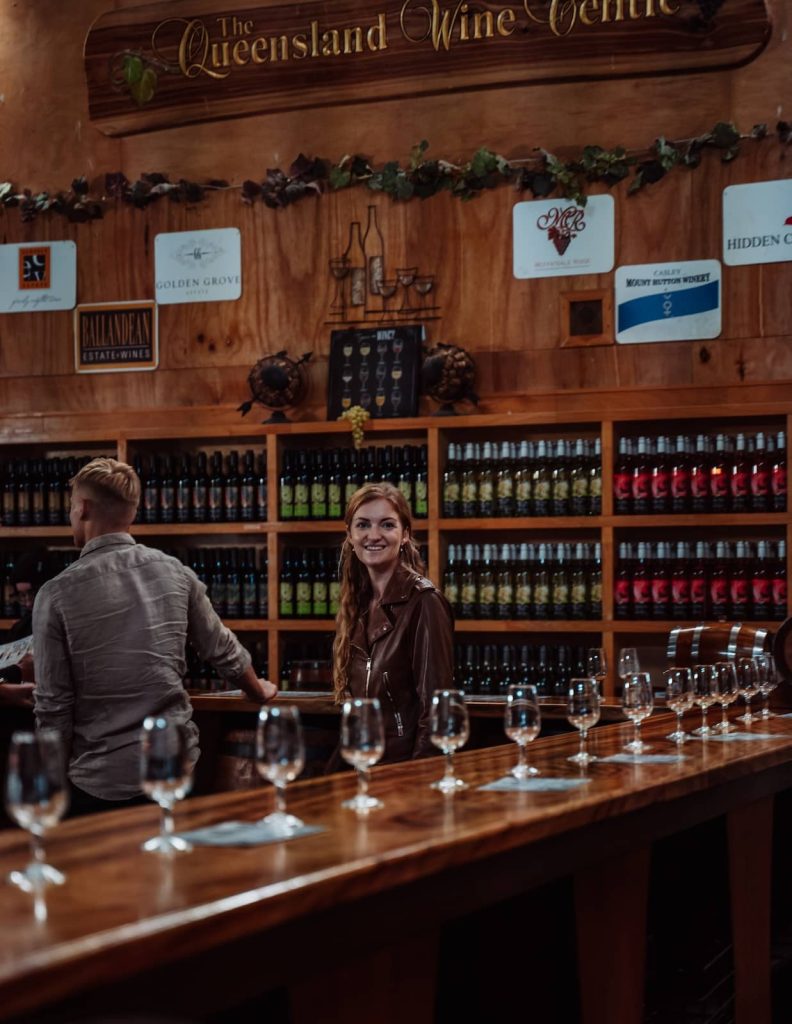 A woman in a brown leather jacket smiles while standing at a long wooden bar inside the Queensland Wine Centre. Rows of empty wine glasses are lined up on the counter, ready for a tasting, with shelves of colorful wine bottles in the background. A man, slightly out of focus, is engaged in conversation at the bar.