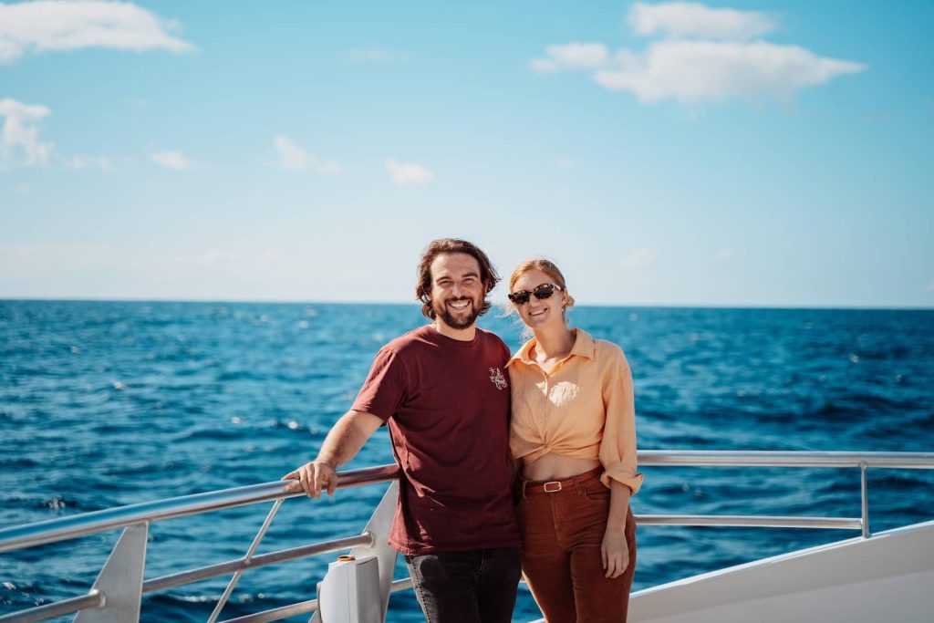 A smiling couple stands on the deck of a boat, leaning against the railing with the vast blue ocean behind them. The man wears a burgundy t-shirt and jeans, while the woman is dressed in a tied-up peach shirt and brown pants with sunglasses. The clear sky and open water suggest a peaceful whale-watching cruise off the Gold Coast.