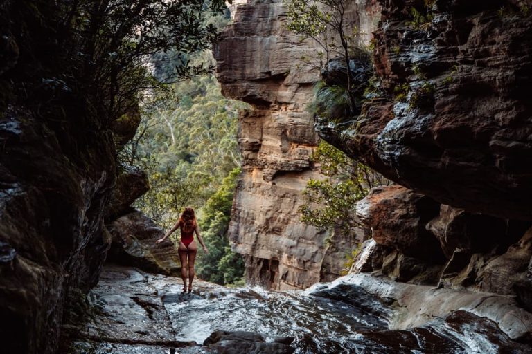 Woman in a red swimsuit standing on wet rock ledge above the secret infinity pool in Leura Cascades, Blue Mountains, surrounded by steep sandstone cliffs and dense green bushland.