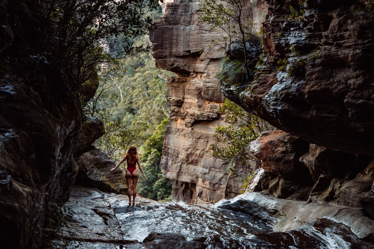 Woman in a red swimsuit standing on wet rock ledge above the secret infinity pool in Leura Cascades, Blue Mountains, surrounded by steep sandstone cliffs and dense green bushland.