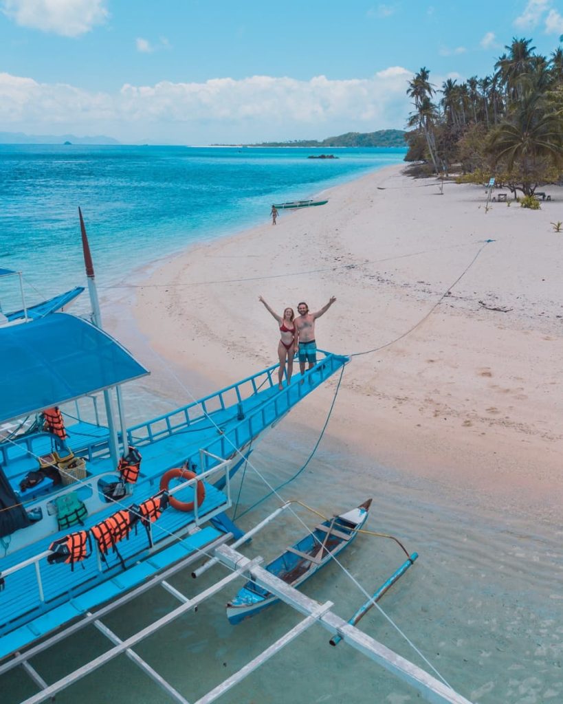 Couple standing on the bow of a blue bangka boat during a Keeloma island hopping tour, one of the most fun things to do between Coron and El Nido, Philippines.
