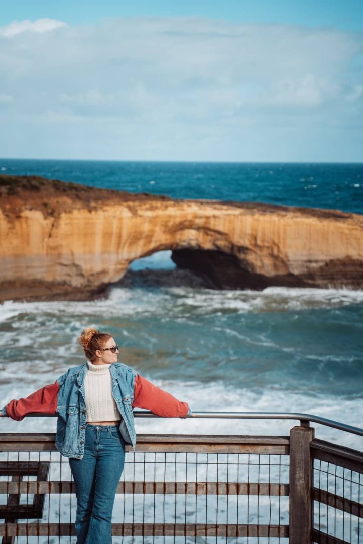 A woman leans on a lookout railing facing the London Arch rock formation along the coast, a famous photo stop on a great ocean road itinerary.