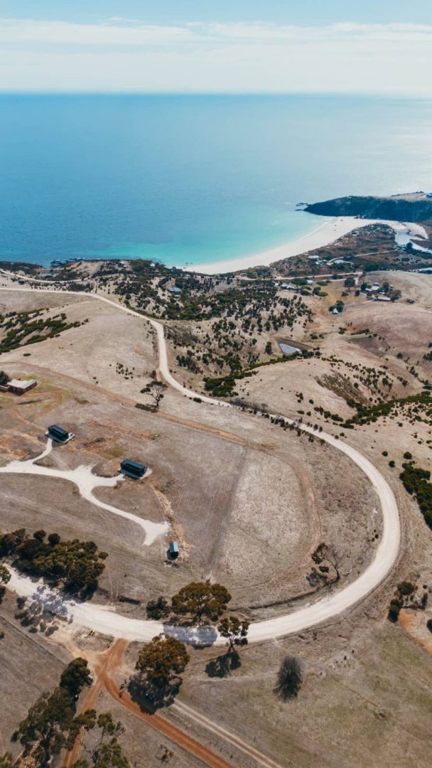 Bird’s-eye view of rolling dry hills and white sandy coastline with bright blue sea on Kangaroo Island, a detour possible during a Melbourne to Darwin road trip.